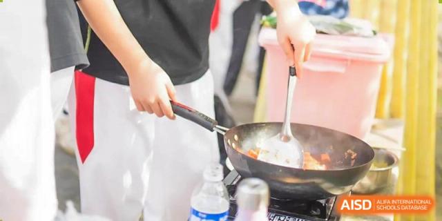 Outdoor cooking activity at BDA School of The High School Affiliated to Renmin University of China, showing a student stirring food in a black non-stick pan with a spatula.