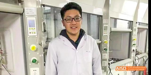 A student from Beijing No.2 Middle School wearing glasses and a white lab coat, standing and smiling in a laboratory or technical workshop environment.