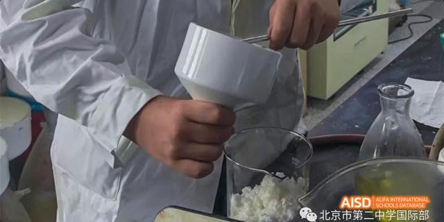 A student from Beijing No.2 Middle School wearing a lab coat, adding a white powdery substance to a beaker or glass container.