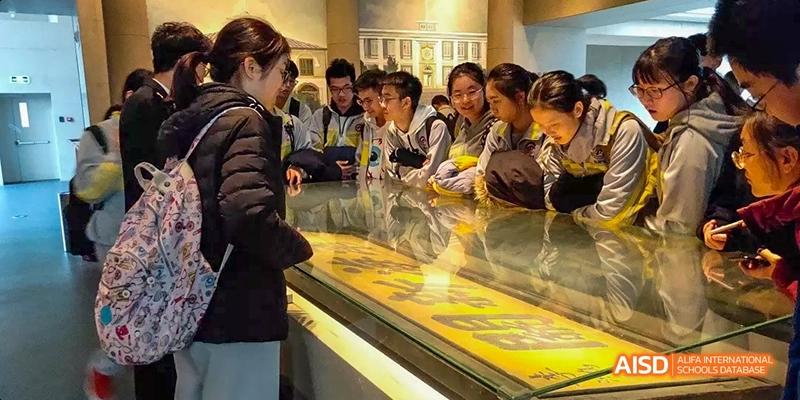 A group of students from Beijing No.2 Middle School gathered around a glass display case inside a museum, looking intently at an exhibit.