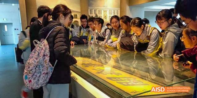 A group of students from Beijing No.2 Middle School gathered around a glass display case inside a museum, looking intently at an exhibit.