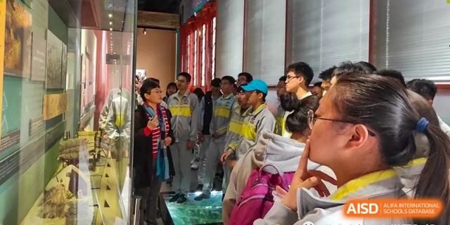 A large group of students from Beijing No.2 Middle School, many wearing grey and yellow uniforms, standing and looking at an exhibit or through a doorway inside a building with red and gold traditional Chinese decor.