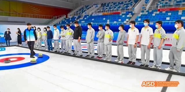 A line of students from Beijing No.2 Middle School in matching light grey and yellow sportswear, standing on the edge of a curling rink, possibly receiving instruction.