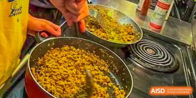 A close-up of a pan on a stove, showing a cooking activity with a dish of ground yellow food being stirred by a student from Beijing No.2 Middle School.