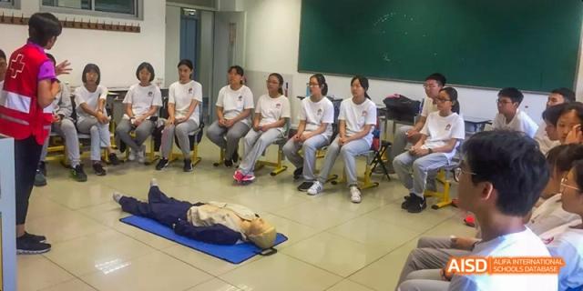 A group of female students from Beijing No.2 Middle School sitting in a semicircle and watching a demonstration with a CPR mannequin lying on a mat.