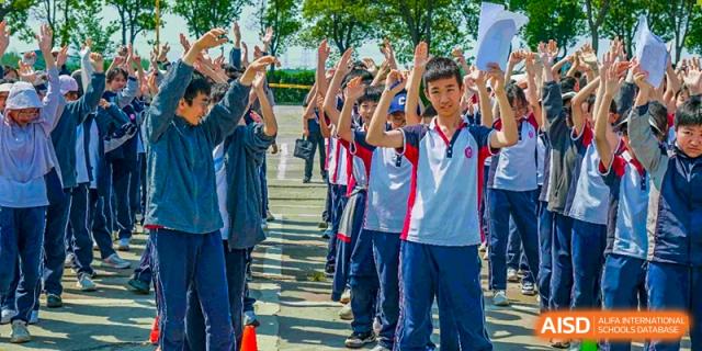 A line of male students from The International School of Rhodes in uniform with their hands raised during an outdoor assembly or activity.