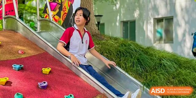 A female student from The International School of Rhodes in uniform sitting on a metal slide in a green outdoor area.