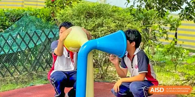 A student from The International School of Rhodes pouring liquid into a large blue funnel structure as part of an outdoor activity.