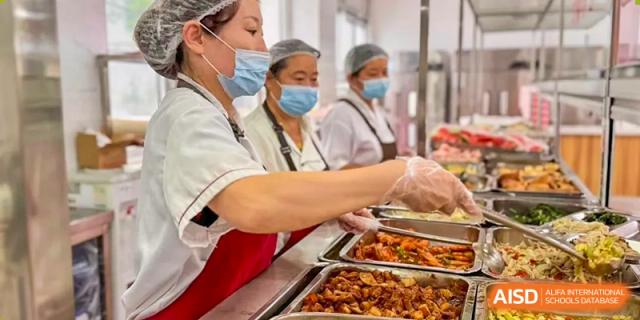Kitchen staff from The International School of Rhodes serving food from metal trays in the school cafeteria.