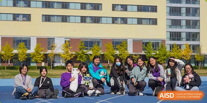 A group of happy students from Tsinghua International School Daoxiang Lake crouching on a blue track and posing for a picture with a small white dog in front of a school building.