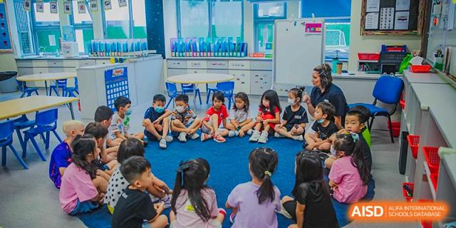 Young students at American International School of Guangzhou (AISG) sit in a circle on the floor, listening to a teacher in a bright classroom setting.