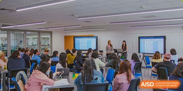 Students at American International School of Guangzhou (AISG) attend a large class or lecture in a modern room with a digital whiteboard display.