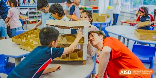 Young students at American International School of Guangzhou (AISG) collaborate with an adult to build a small wooden structure or model.