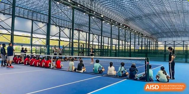 Students sitting on the sideline of a large indoor blue sports court with a high ceiling, watching a padel or tennis game at the American International School of Guangzhou (AISG).