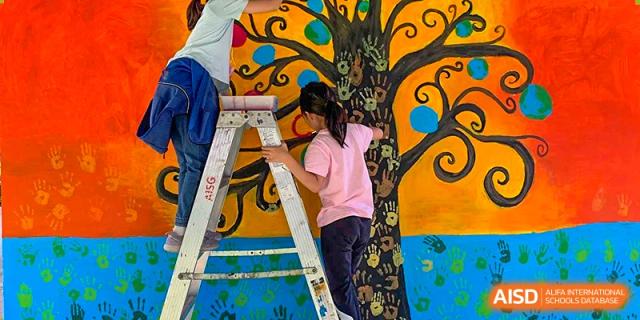 A female student standing on a ladder and painting a large, colorful mural or scenic art piece in a school facility at the American International School of Guangzhou (AISG).