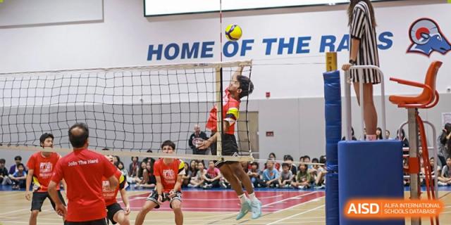 A student athlete from American International School of Guangzhou (AISG) spikes a volleyball over the net during a game, with the text "HOME OF THE" visible on the wall in the background.