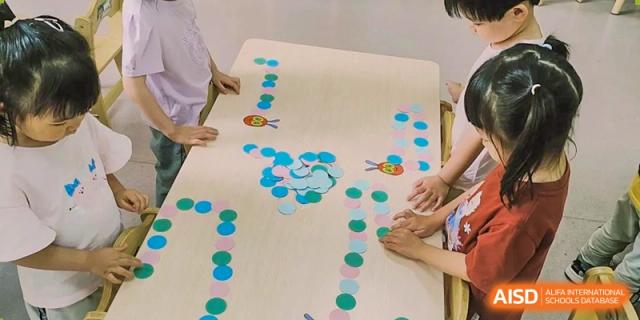 Several people gathered around a large wooden table with small colorful paper circles arranged in paths and designs, engaged in a table activity at Britannia International School Guangzhou.