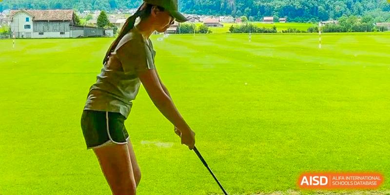 A young person outdoors practicing a golf swing on a bright green course, with a grassy field and trees in the background, at Britannia International School Guangzhou.