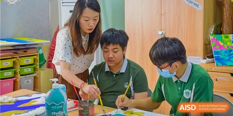 A teacher assisting two students with a painting activity in a classroom, where they are using yellow and green paints, at Britannia International School Guangzhou.