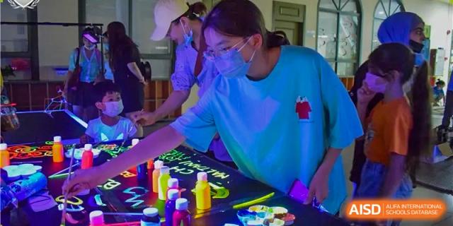 A student wearing a face mask and glasses painting on a black surface under fluorescent lighting with colorful paints and materials at Britannia International School Guangzhou.