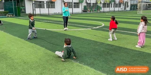 A young child sitting on a green sports field facing a female adult standing nearby, during an outdoor activity at Britannia International School Guangzhou.