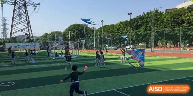 A student throwing a ball on a green sports field with a large blue kite flying in the background at Britannia International School Guangzhou.