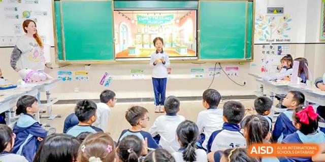 A teacher giving a presentation or lesson in a brightly lit classroom using a large screen to an audience of students at the Affiliated School of JNU for Hong Kong & Macao Students.