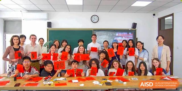 A large group of students sitting with a male teacher, holding up red envelopes and certificates in a classroom celebration at Beijing Academy International Department (BAID).
