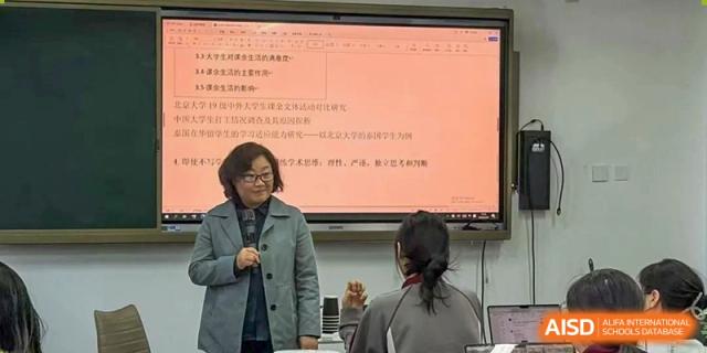 A teacher stands in front of a digital screen, giving a lecture to a student in the foreground and a class full of students at Beijing Academy International Department (BAID).