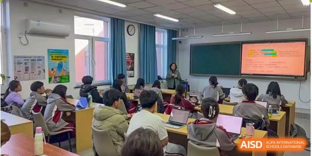 A teacher instructs students who are seated at desks and working on laptops in a bright classroom at Beijing Academy International Department (BAID).