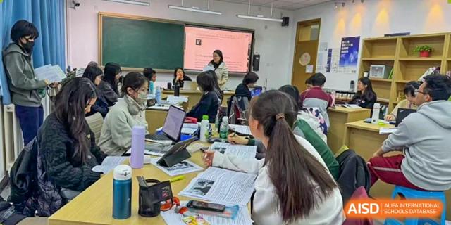 Students working in small groups on various tasks, with a teacher visible near a screen in the background at Beijing Academy International Department (BAID).
