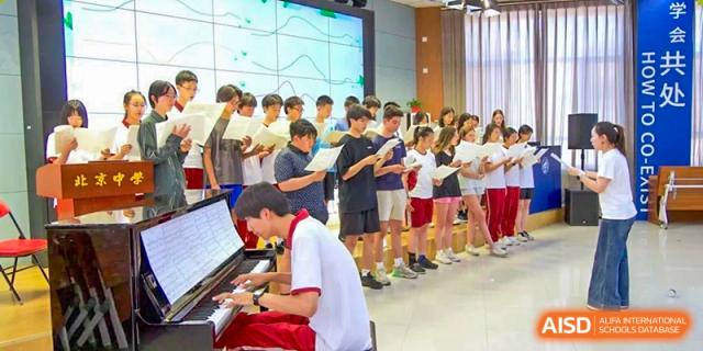 A group of students singing in a chorus, accompanied by a student playing the piano, in a well-lit modern hall at Beijing Academy International Department (BAID).