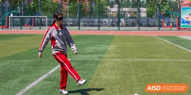 A female student in a red and white uniform kicking a ball on a green sports field during practice or a game at Beijing Academy International Department (BAID).