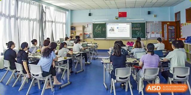 An image showing the backs of students seated at desks in a clean, brightly lit classroom, looking toward the front where a screen and chalkboard are visible, at Beijing SMIC Private School.
