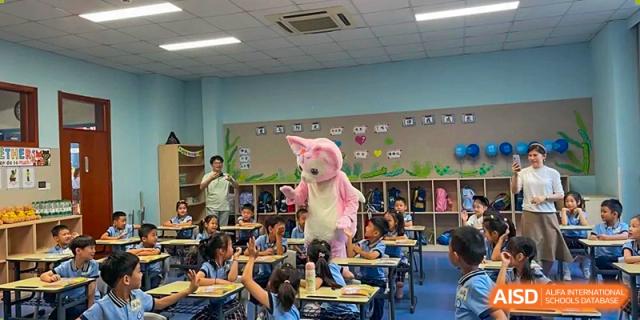 A group of young students in a classroom interacting enthusiastically with a person dressed in a large pink rabbit costume, at Beijing SMIC Private School.
