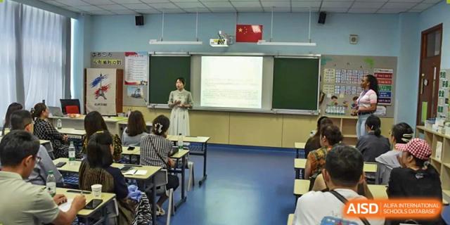 A female teacher standing at the front of a classroom, instructing students with a presentation visible on a projection screen, at Beijing SMIC Private School.