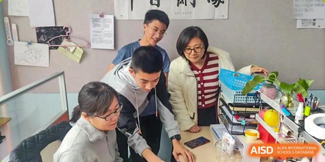 A teacher or adult guiding a group of smiling students as they collaborate on a project on a table, at Beijing SMIC Private School.