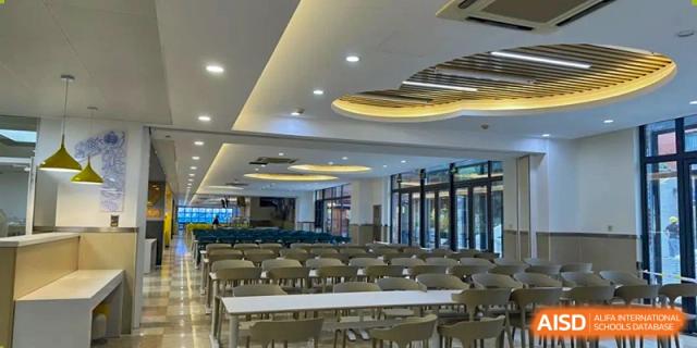 An interior view of the large, modern school cafeteria or dining hall with many tables and chairs under unique circular lighting, at Beijing SMIC Private School.