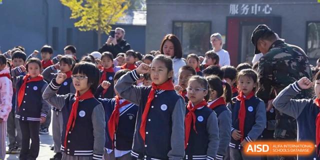 A group of young students in uniform with red scarves standing outdoors in formation, with a female teacher observing them, at Beijing SMIC Private School.