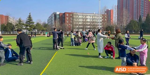 A group of students and parents participating in a fitness or warm-up activity on a green turf field with a large red brick school building in the background, at Beijing SMIC Private School.