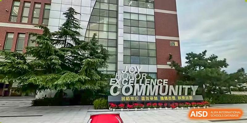 A close-up of a modern school entrance facade with large windows and a sign featuring the words "JOY," "EXCELLENCE," and "COMMUNITY," at Beijing SMIC Private School.