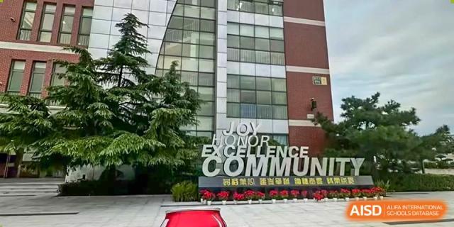 A close-up of a modern school entrance facade with large windows and a sign featuring the words "JOY," "EXCELLENCE," and "COMMUNITY," at Beijing SMIC Private School.