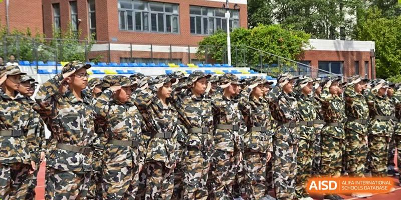 A group of students wearing camouflage uniforms standing in formation during a military training or school drill, with a stadium stand visible in the background, at Beijing SMIC Private School.