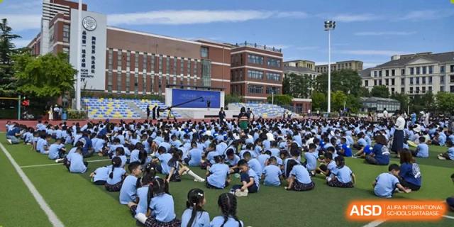 A large gathering of students in blue uniforms seated on a sports field, watching a performance or event on a stage in front of the main school building, at Beijing SMIC Private School.