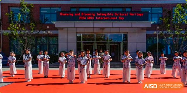 A group of female students performing a traditional dance in white and pink costumes in front of the school building entrance, with a banner overhead, at Beijing SMIC Private School.