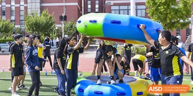 Students participating in a team-building or sports activity, standing on an inflatable blue platform while maneuvering a large, colorful object, at Beijing SMIC Private School.