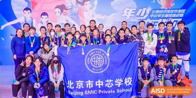 A large group of students and teachers smiling and posing with medals, holding a blue banner that displays the school logo and name, at Beijing SMIC Private School.