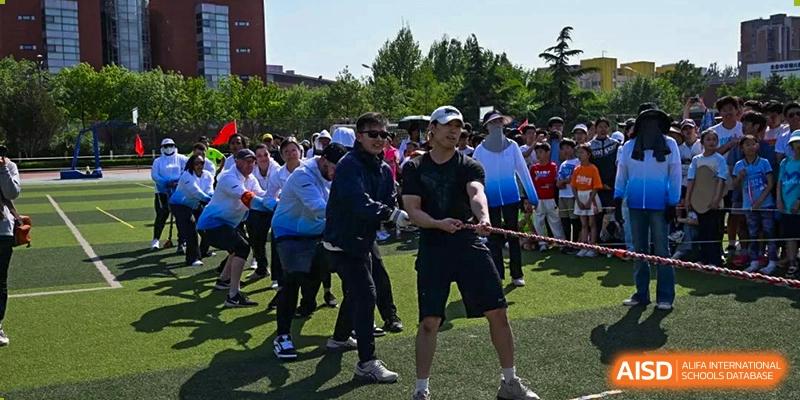 Adults smiling and participating in a team relay race or field day activity, pulling on a rope or similar equipment on a sports field, at Beijing SMIC Private School.