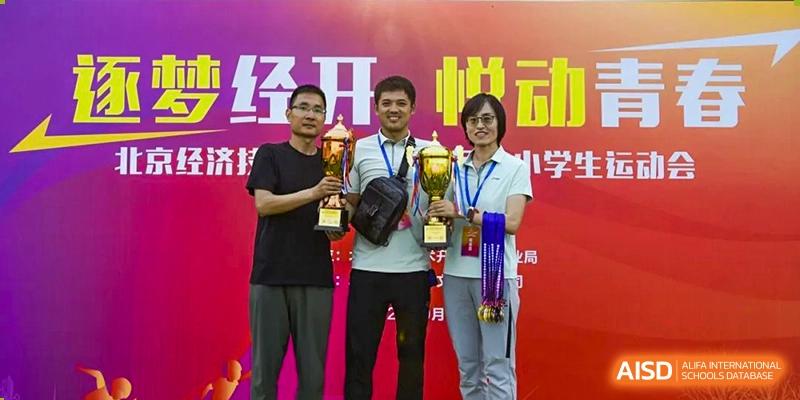 Two male and one female teacher or staff member smiling and proudly holding large gold trophies and awards during a school ceremony, at Beijing SMIC Private School.
