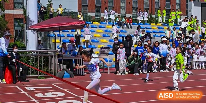 A young girl participating in an outdoor track and field event, running in a sack race in front of seated spectators in the stands, at Beijing SMIC Private School.
